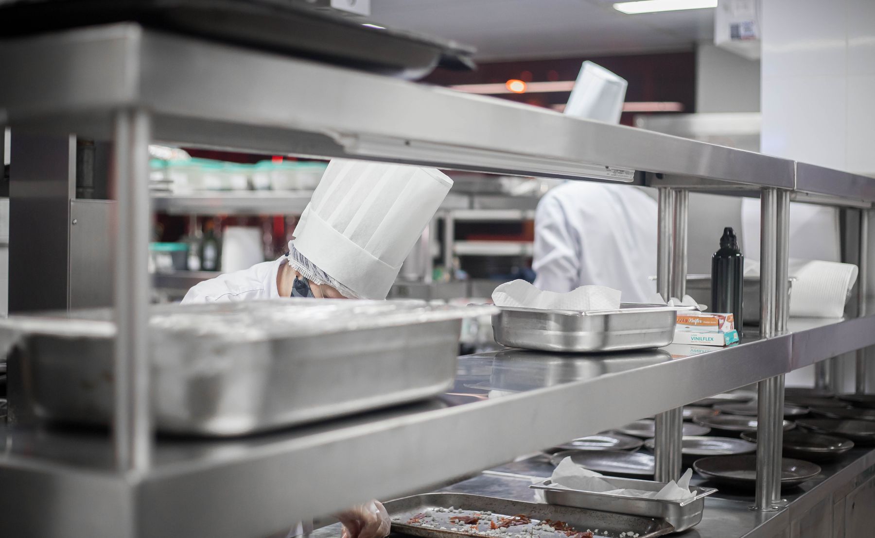 Cooks preparing food in a stainless steel commercial kitchen with overhead shelving